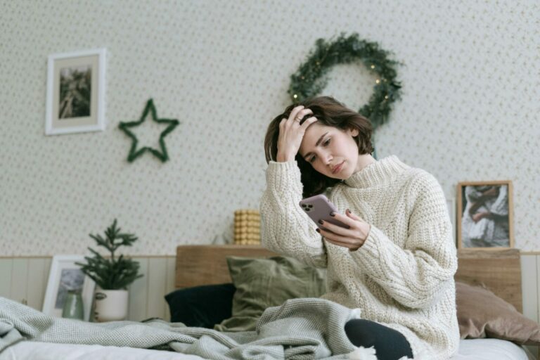 Woman in knitted sweater using smartphone while relaxing in a cozy bedroom.
